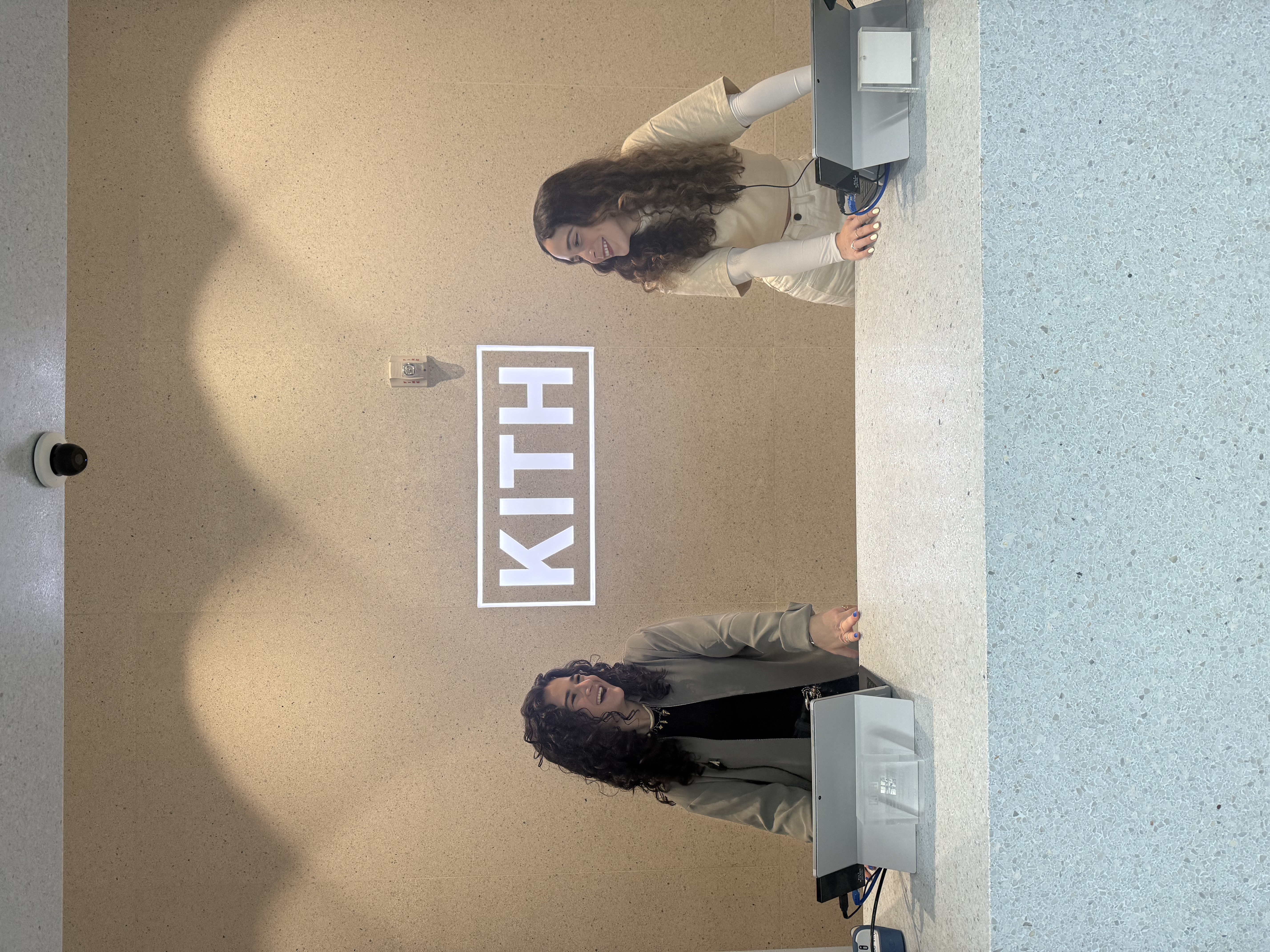 Two Kith Miami Beach team members smiling behind the store counter beneath the illuminated Kith logo